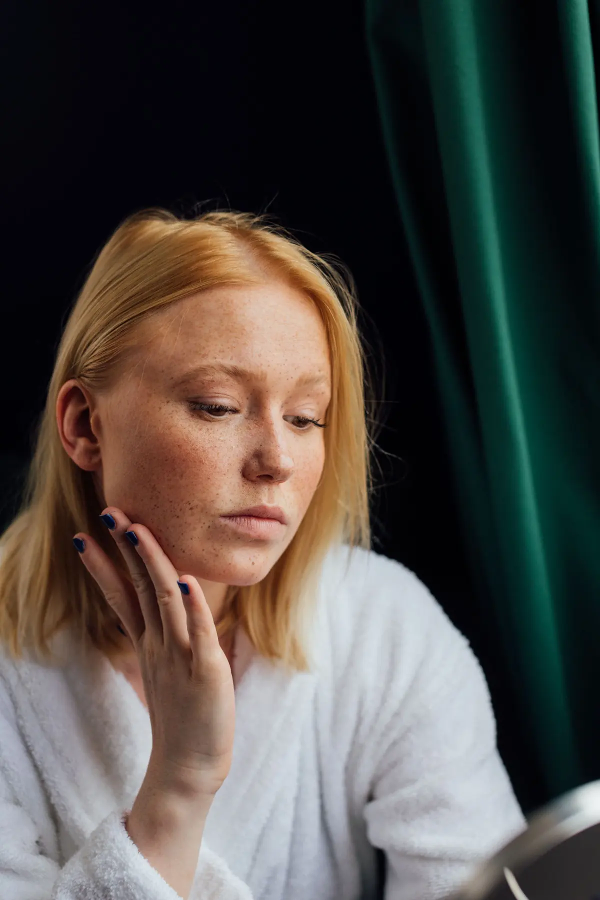 Руда жінка в білому халаті торкається щоки й уважно роздивляється шкіру
